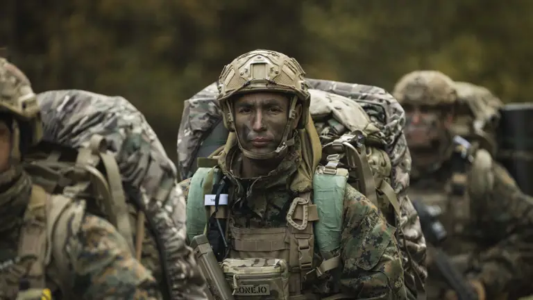 Soldiers in camouflage gear and helmets march through a forest, displaying determination. They carry large backpacks and rifles, conveying focus and readiness.