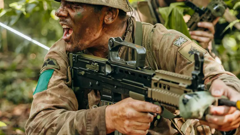 Soldier in camouflage gear aiming a rifle while crouching in dense jungle foliage during a tactical operation.
