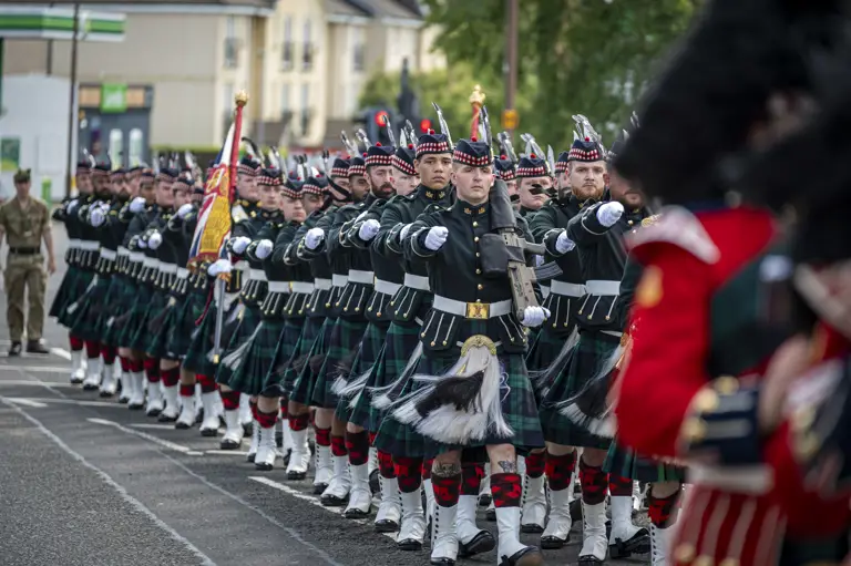 Men wearing black tunics and green and blue kilts march along a street. They are carrying rifles.
