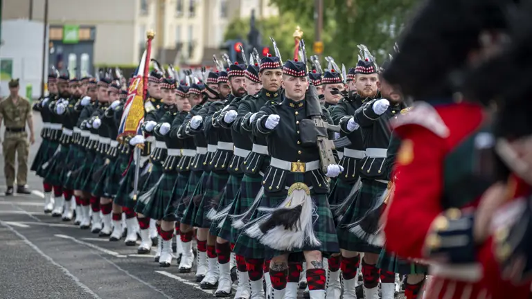 Men wearing black tunics and green and blue kilts march along a street. They are carrying rifles.