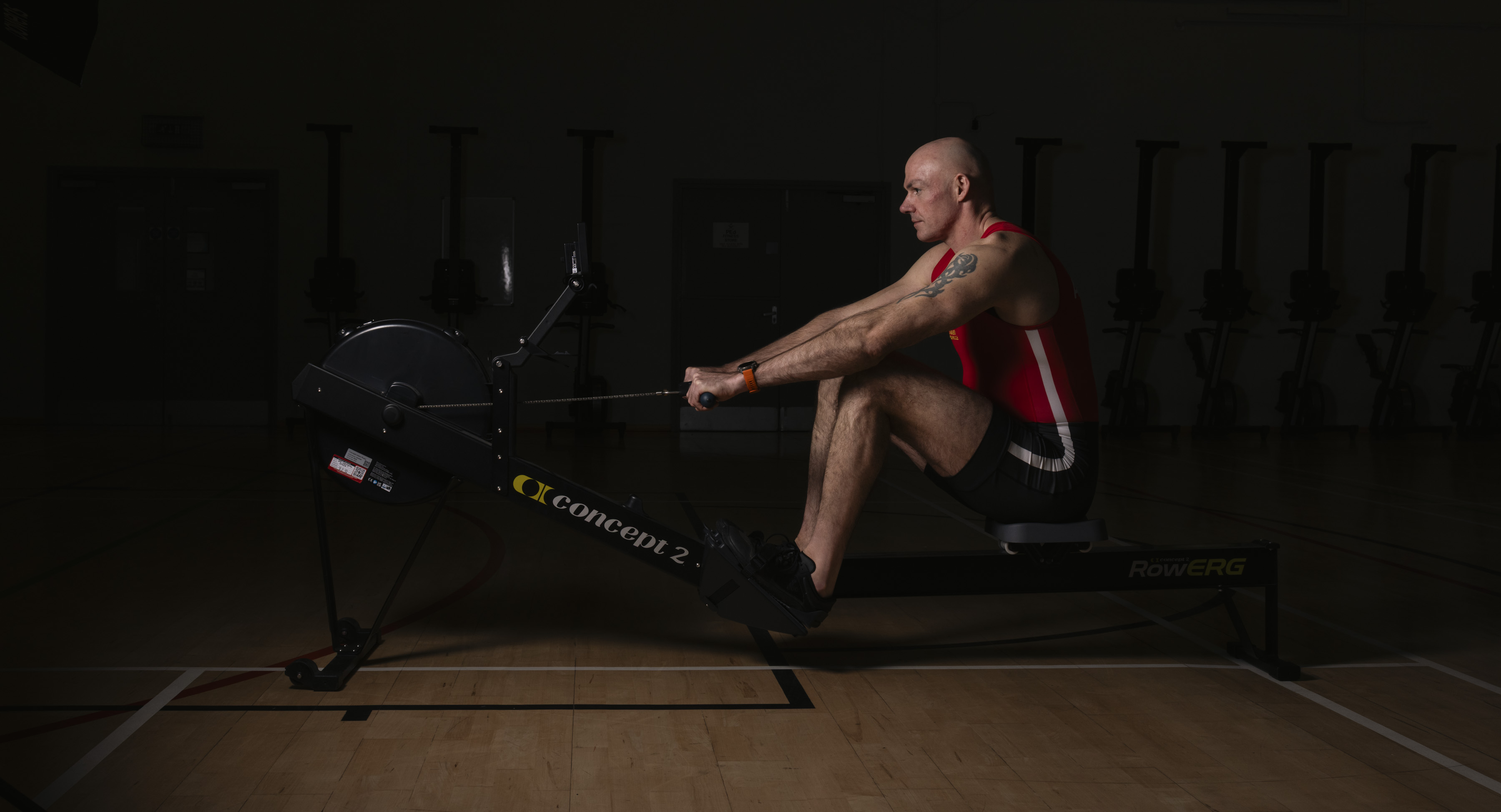 A man in a red top and black shorts uses an indoor rowing machine.