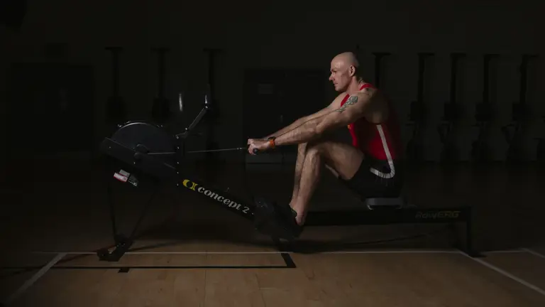 A man in a red top and black shorts uses an indoor rowing machine.