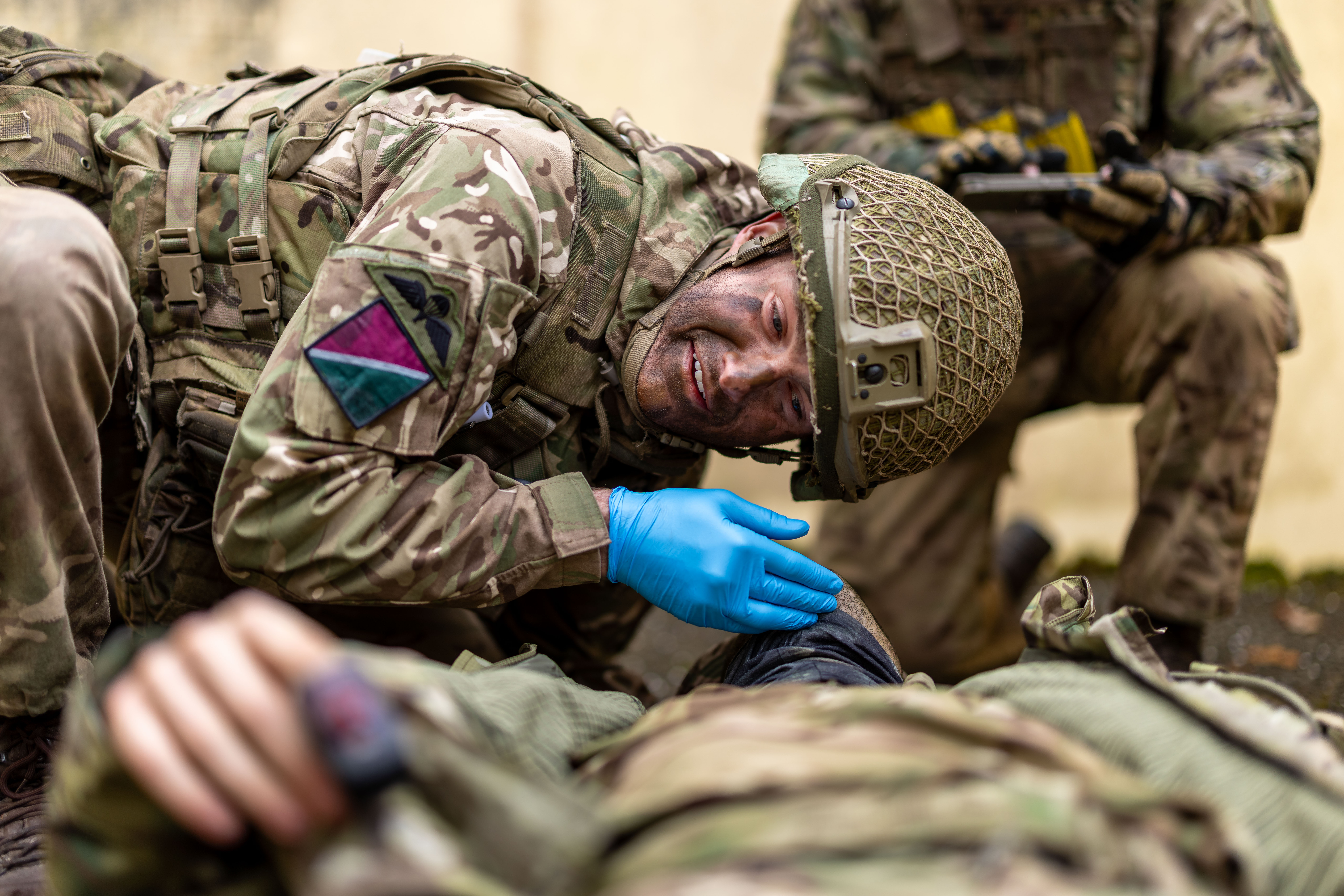 A person in camouflage military gear, wearing blue gloves and a helmet, is attending to another individual.