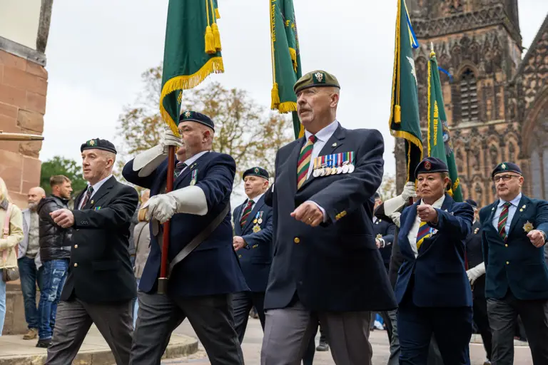 Veterans in suits, berets and medals march in front of the cathedral holding colours flags.