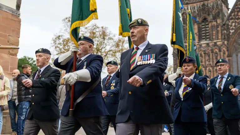 Veterans in suits, berets and medals march in front of the cathedral holding colours flags.