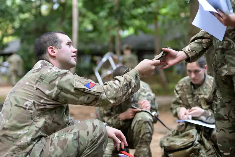 An Army clerk in camouflage uniform issues local currency to soldiers, also in camouflage uniform who have newly arrived abroad in support of Operation Gritrock.