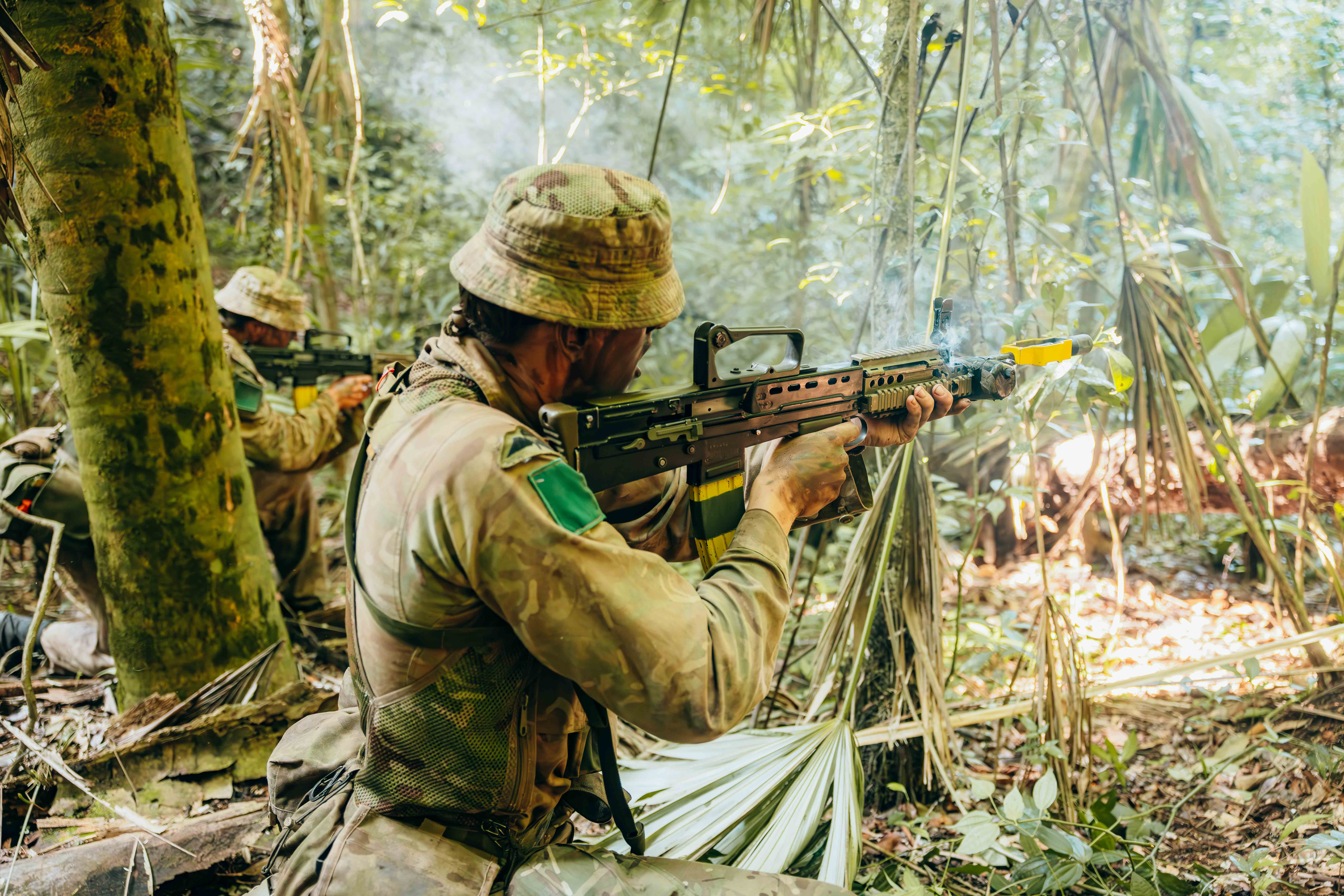 Soldier in camouflage gear aiming a rifle while crouched in dense jungle foliage during a military exercise.