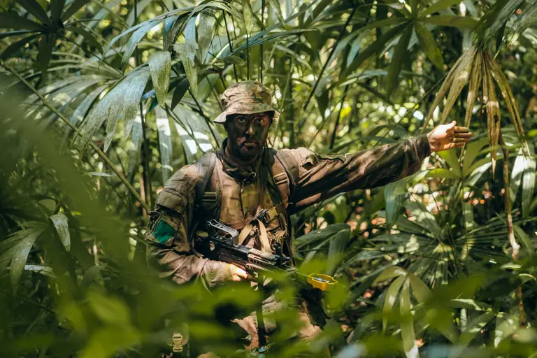Soldier in full camouflage gear signaling with his arm while standing in dense jungle foliage.