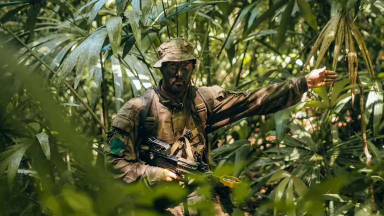 Soldier in full camouflage gear signaling with his arm while standing in dense jungle foliage.