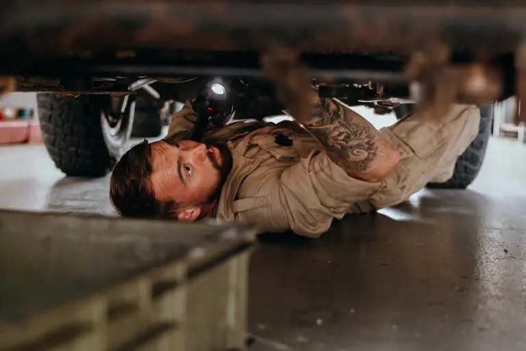 Mechanic in beige coveralls inspecting the underside of a vehicle using a flashlight in a garage.