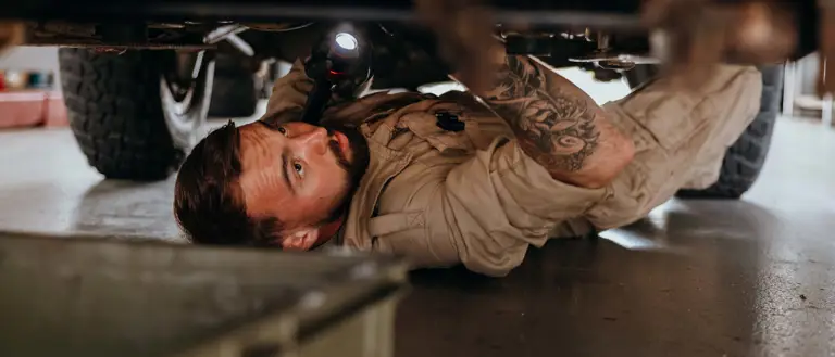 Mechanic in beige coveralls inspecting the underside of a vehicle using a flashlight in a garage.