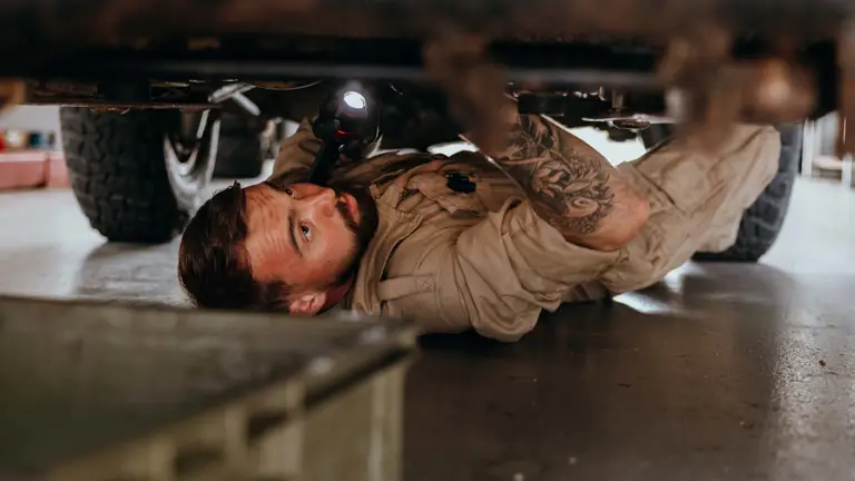 Mechanic in beige coveralls inspecting the underside of a vehicle using a flashlight in a garage.