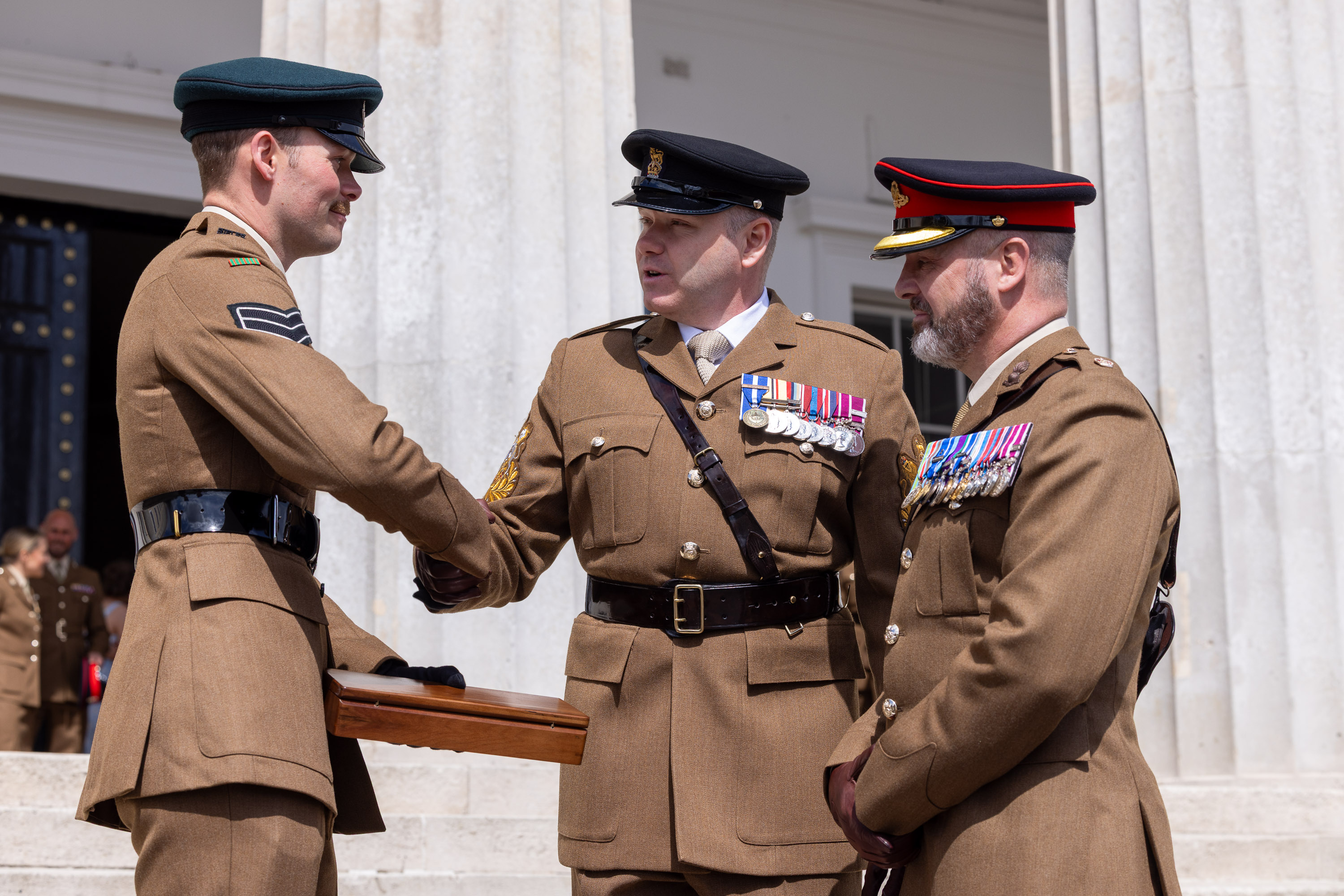 Three men in brown uniforms, two decorated with medals are stood shaking hands outside of a stone building.