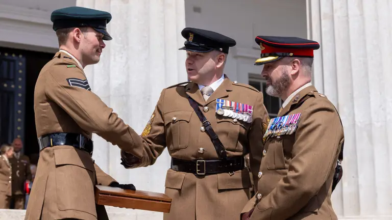 Three men in brown uniforms, two decorated with medals are stood shaking hands outside of a stone building.