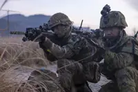 One British soldier and one Japanese soldier, both in camouflage uniform holding issued rifles while kneeing on the ground focusing on their out of frame target on a training area in Japan.