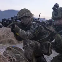 One British soldier and one Japanese soldier, both in camouflage uniform holding issued rifles while kneeing on the ground focusing on their out of frame target on a training area in Japan.