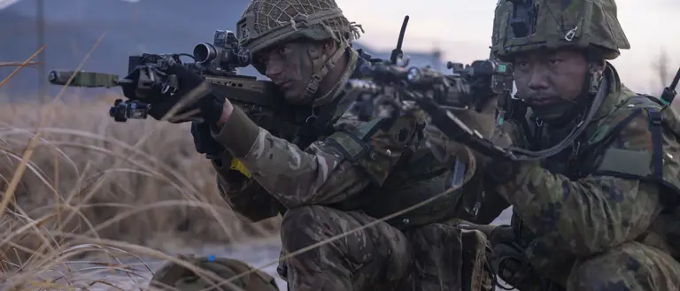 One British soldier and one Japanese soldier, both in camouflage uniform holding issued rifles while kneeing on the ground focusing on their out of frame target on a training area in Japan.