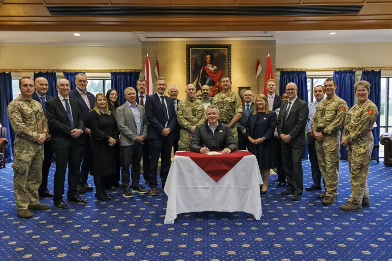 Group of military personnel and civilians posing around a seated man signing a document in a formal room with flags.