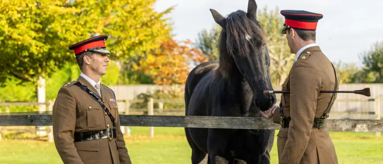 Two British Army officers in khaki uniforms, are standing either side of a black horse that is leaning over a fence.