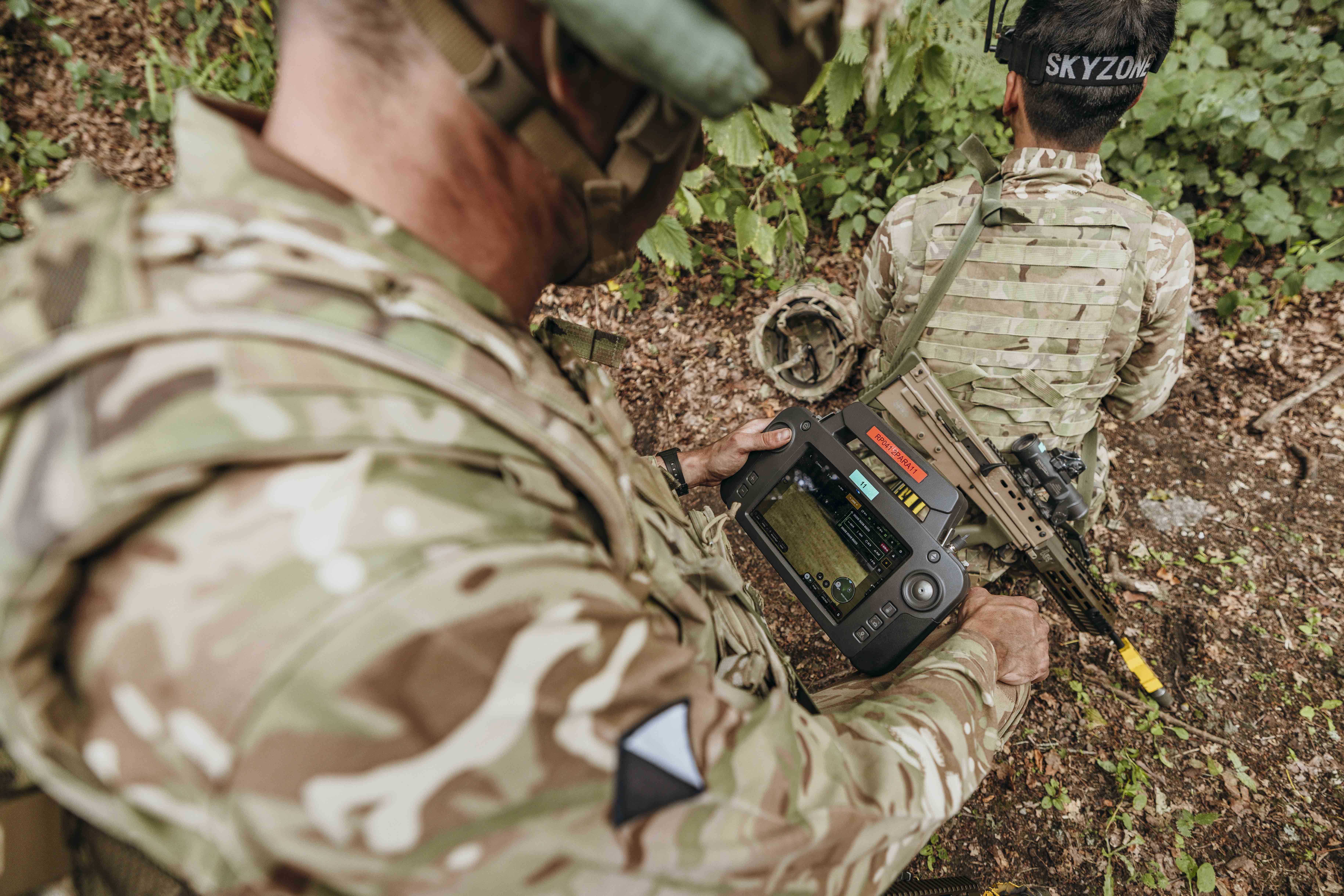 Soldier in camouflage gear operating a handheld tactical device while another soldier kneels nearby in a wooded area.