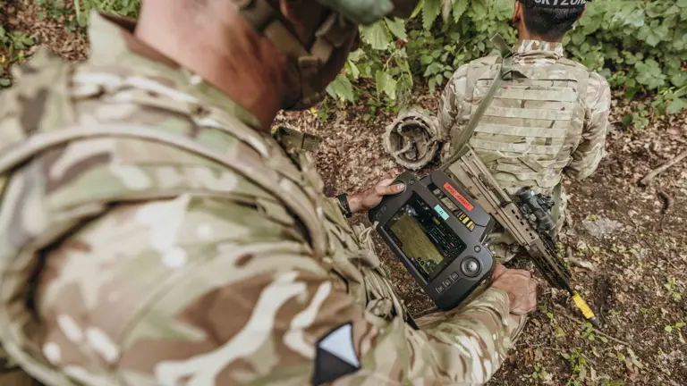 Soldier in camouflage gear operating a handheld tactical device while another soldier kneels nearby in a wooded area.