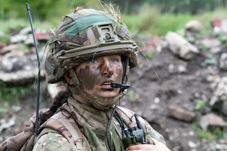 Female officer cadet seen in camouflage uniform and camo cream on her face speaking into a radio