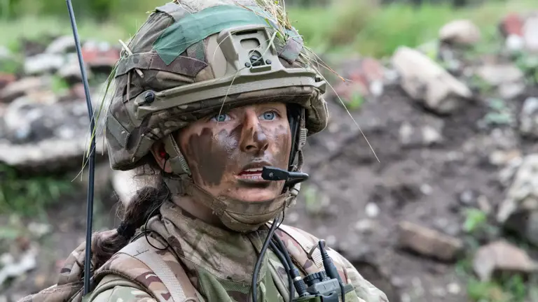 Female officer cadet seen in camouflage uniform and camo cream on her face speaking into a radio