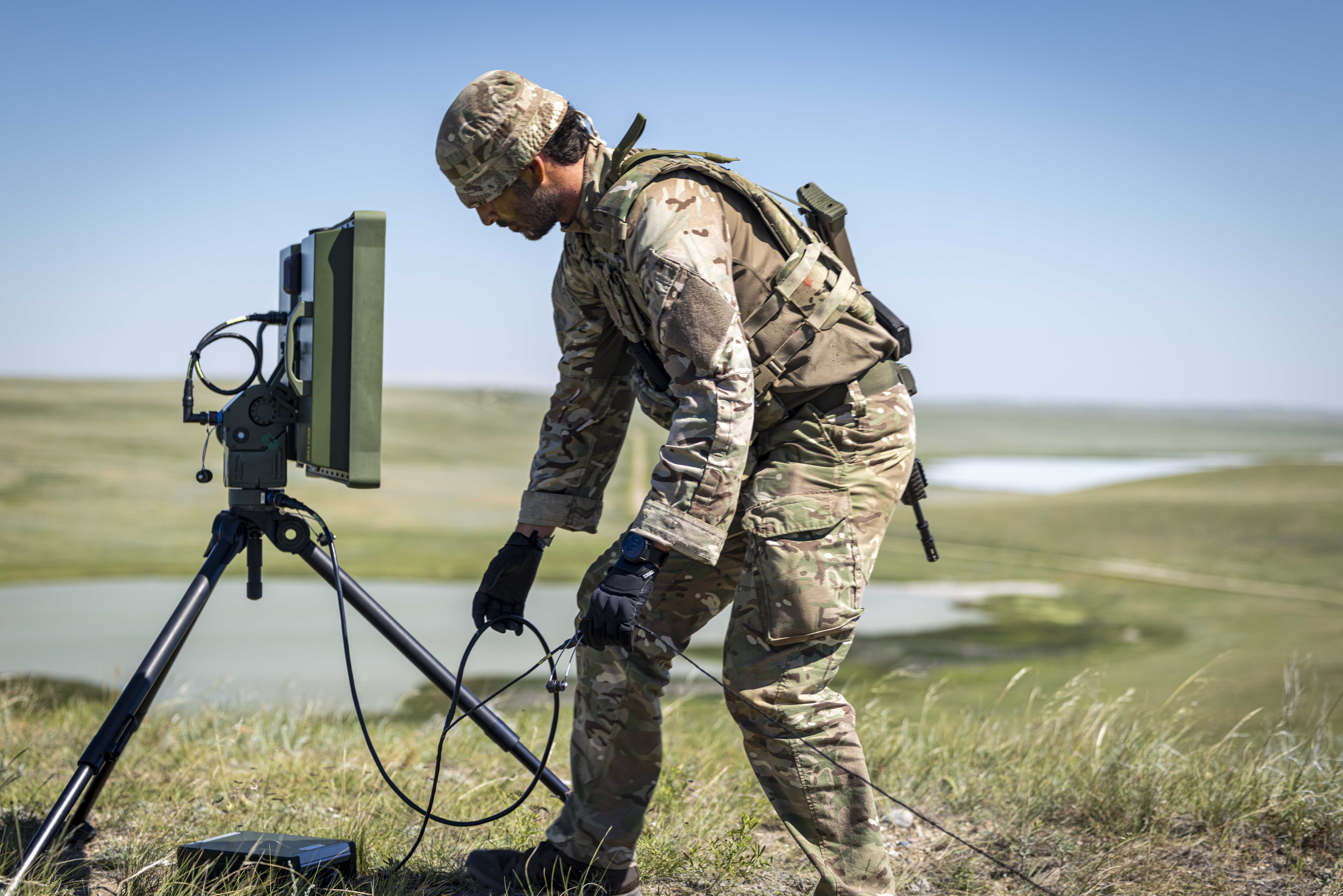 A person in camouflage military attire is kneeling beside a portable radar system on a tripod in an open grassy field. The landscape features distant rolling hills and a body of water. The person is holding a small device connected to the radar system.