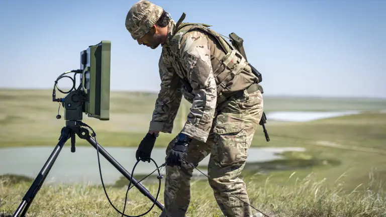 A person in camouflage military attire is kneeling beside a portable radar system on a tripod in an open grassy field. The landscape features distant rolling hills and a body of water. The person is holding a small device connected to the radar system.