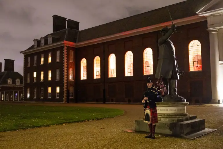 A soldier in traditional Scottish uniform stands guard beside a statue whilst holding a bagpipe.