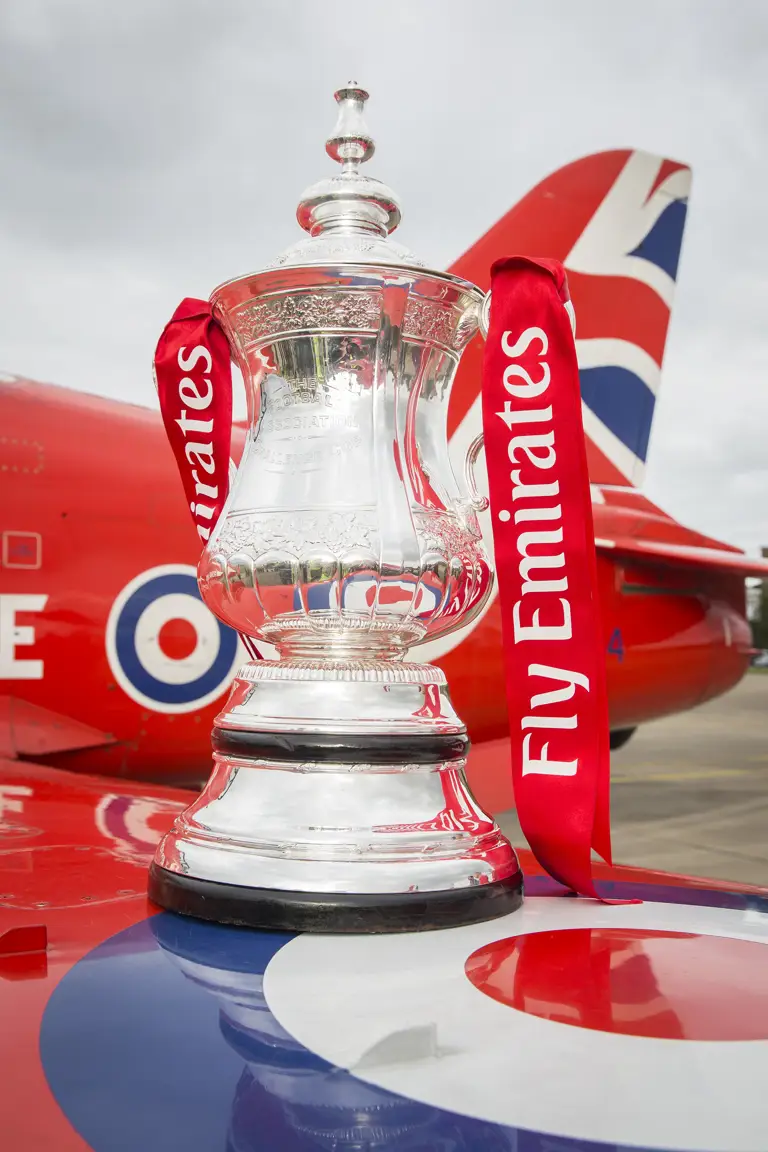 The image shows the FA Cup trophy with red ribbons that say