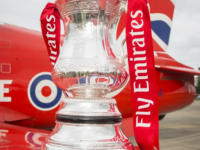 The image shows the FA Cup trophy with red ribbons that say