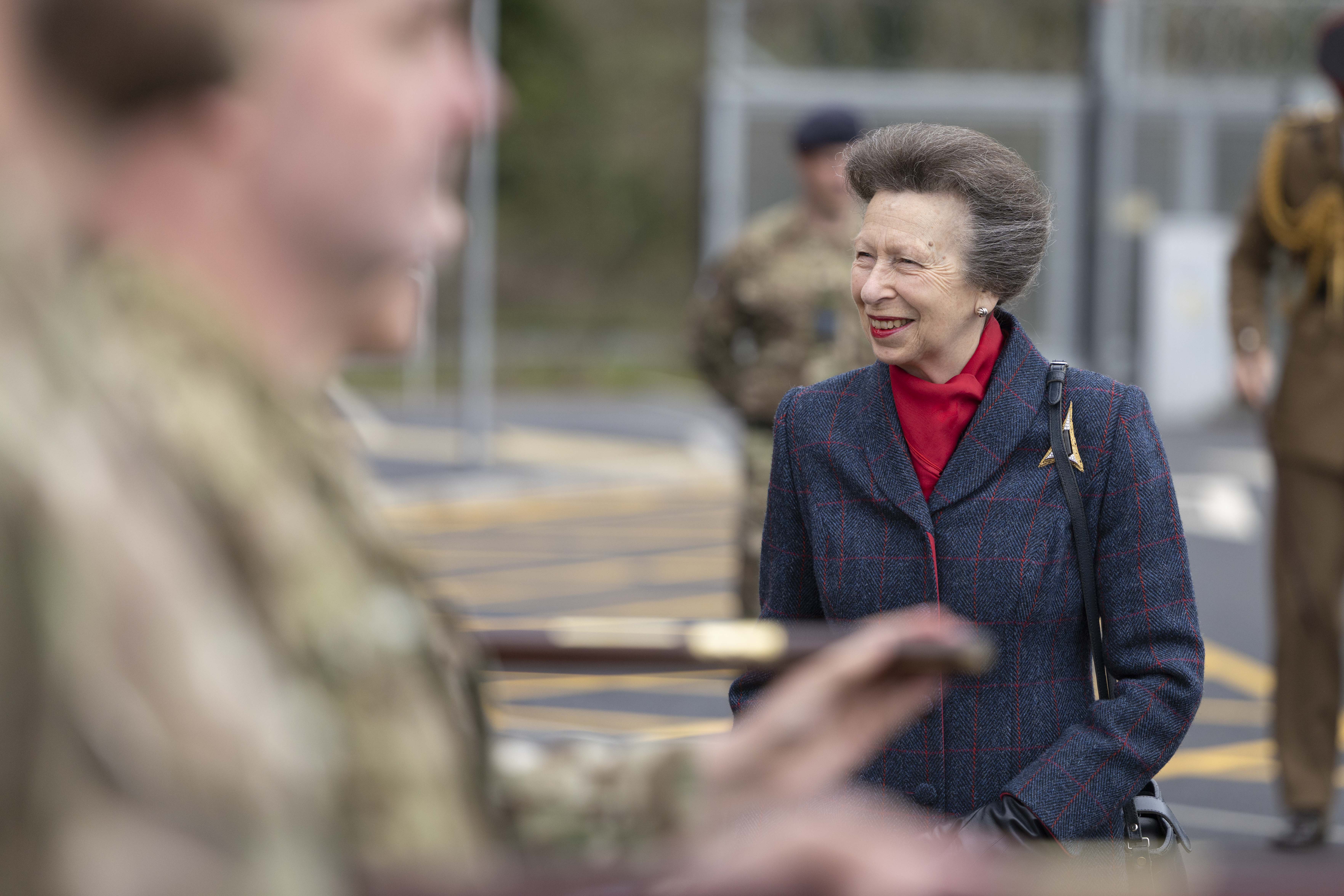 A person wearing a red plaid coat stands among several uniformed military personnel. They are engaged in an outdoor ceremonial event, with the military personnel holding rifles.
