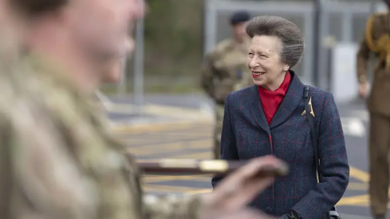 A person wearing a red plaid coat stands among several uniformed military personnel. They are engaged in an outdoor ceremonial event, with the military personnel holding rifles.