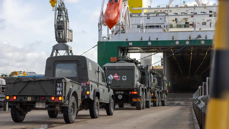 Green military vehicles drive onto a white and green vessel.