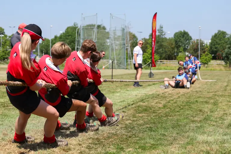 A group of individuals in red shirts are participating in a tug-of-war against another team in blue shirts on a grassy field. An observer stands nearby watching the event. A tall flag with text is seen in the background, and the sky is clear and sunny.