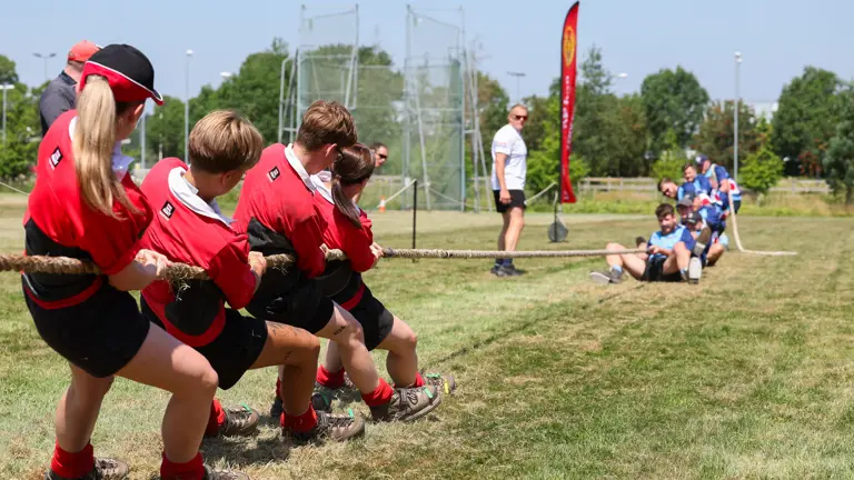 A group of individuals in red shirts are participating in a tug-of-war against another team in blue shirts on a grassy field. An observer stands nearby watching the event. A tall flag with text is seen in the background, and the sky is clear and sunny.