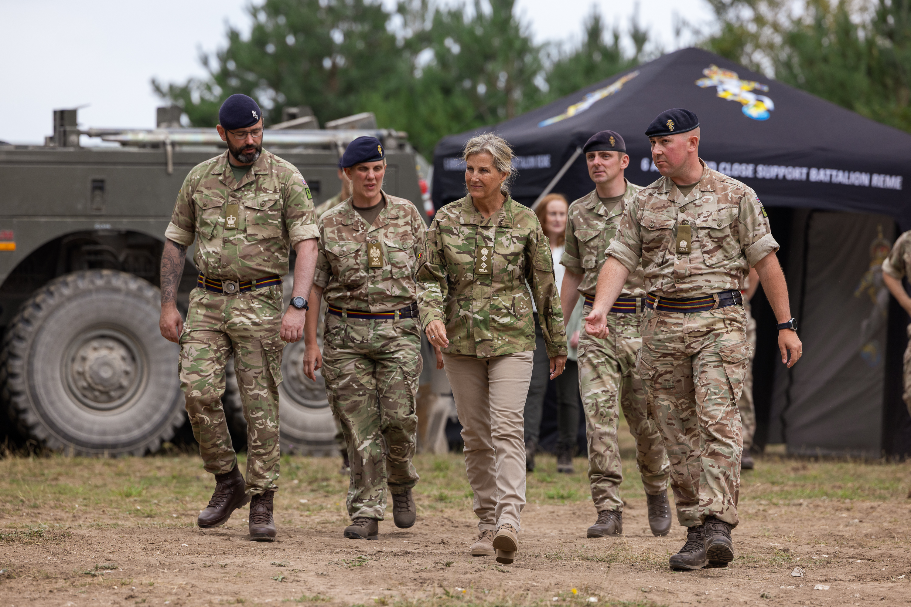 The Duchess of Edinburgh is seen wearing trousers and a camouflage print jacket stood walking outside with two soldiers in camouflage uniform on either side of her.