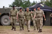 The Duchess of Edinburgh is seen wearing trousers and a camouflage print jacket stood walking outside with two soldiers in camouflage uniform on either side of her.