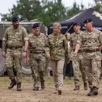 The Duchess of Edinburgh is seen wearing trousers and a camouflage print jacket stood walking outside with two soldiers in camouflage uniform on either side of her.