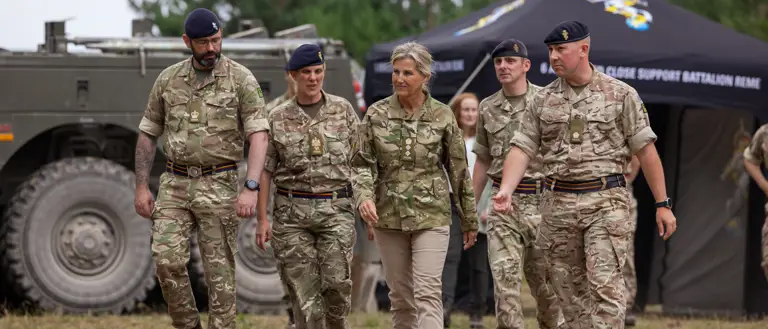 The Duchess of Edinburgh is seen wearing trousers and a camouflage print jacket stood walking outside with two soldiers in camouflage uniform on either side of her.