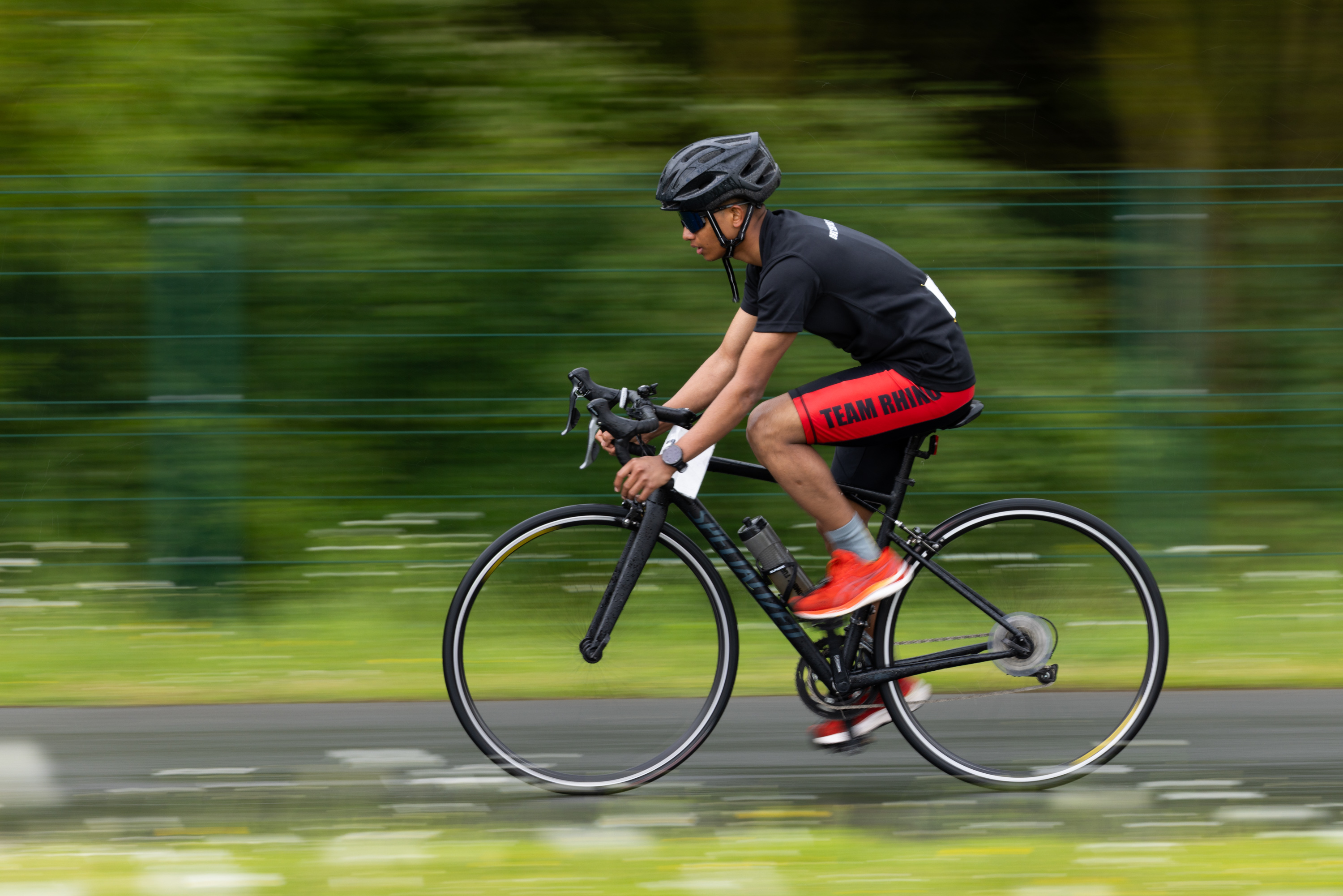 Cyclist in red shorts and black helmet riding fast on a road bike, motion blur emphasizes speed. Background: green foliage and grass.