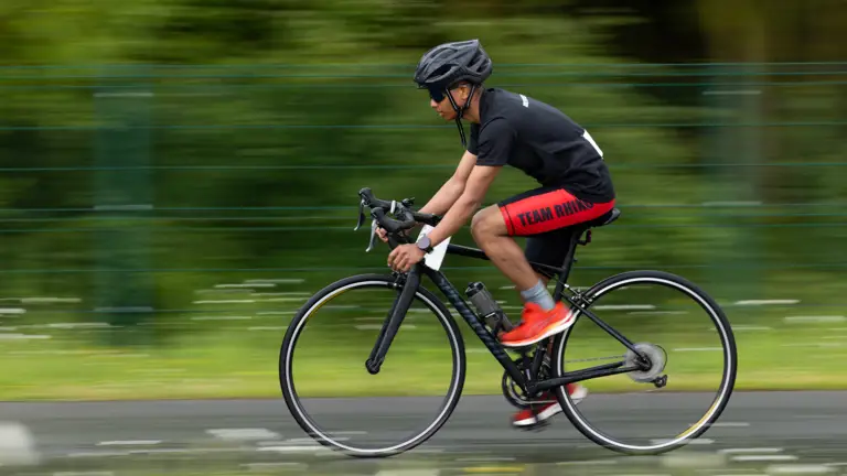 Cyclist in red shorts and black helmet riding fast on a road bike, motion blur emphasizes speed. Background: green foliage and grass.