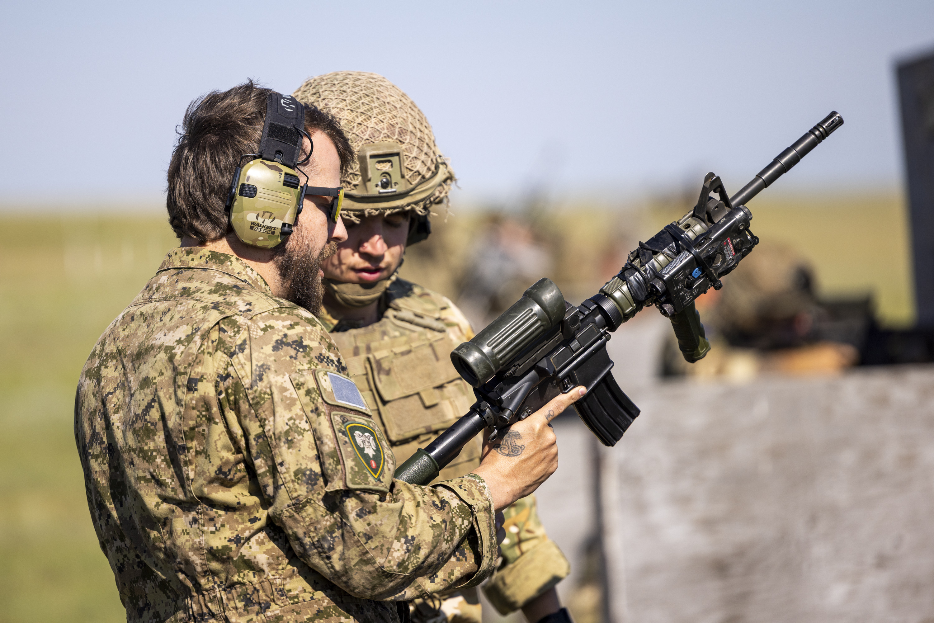 Two individuals in military camouflage are in a grassy field with a wooden gate in the foreground. One is wearing a maroon beret, standing beside the other, who is aiming a rifle toward the distance. The landscape is open and clear under a bright blue sky.
