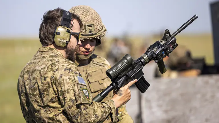 Two individuals in military camouflage are in a grassy field with a wooden gate in the foreground. One is wearing a maroon beret, standing beside the other, who is aiming a rifle toward the distance. The landscape is open and clear under a bright blue sky.