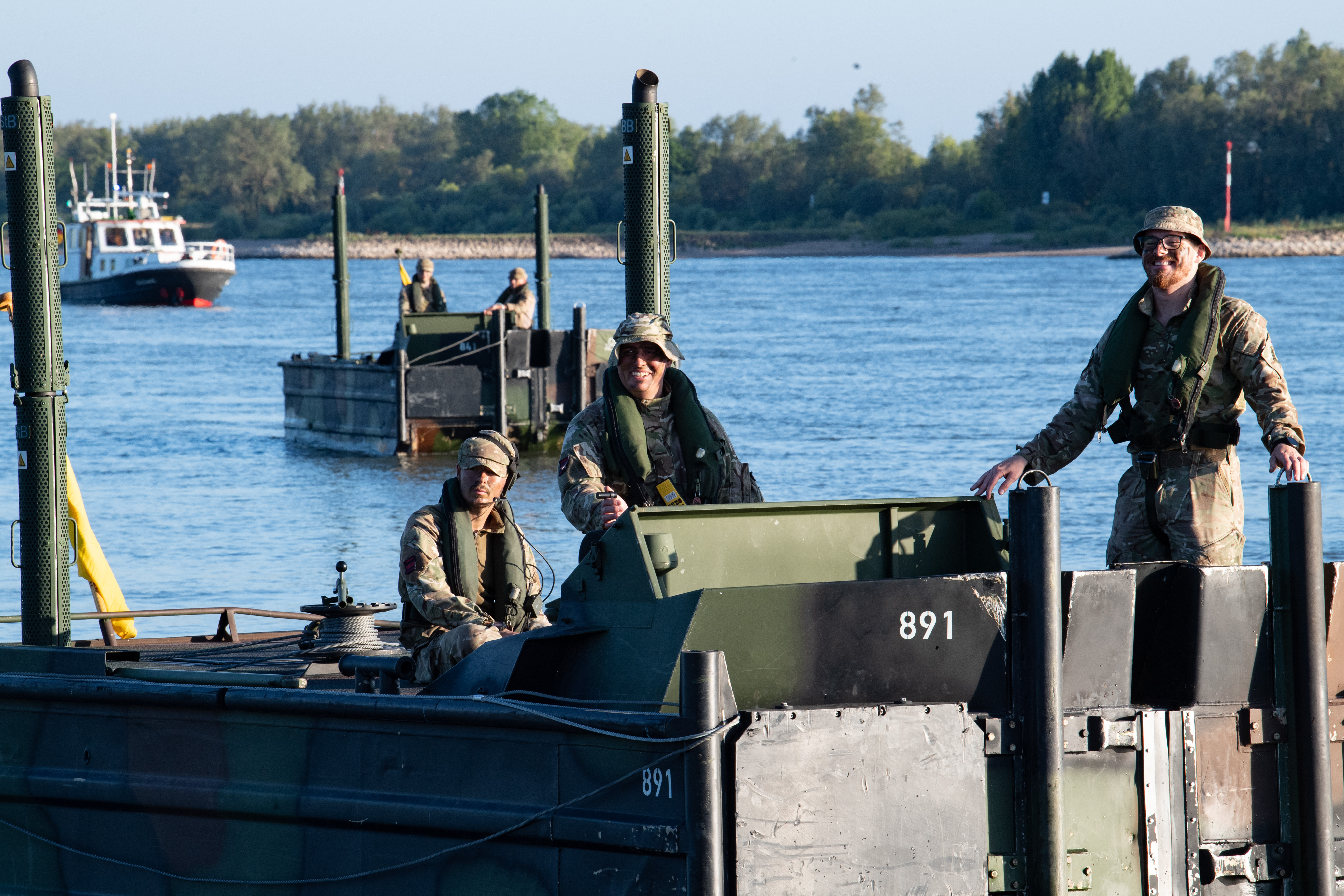 Three soldiers in camouflage uniforms and life vests operate a military boat numbered 891 on a calm river.