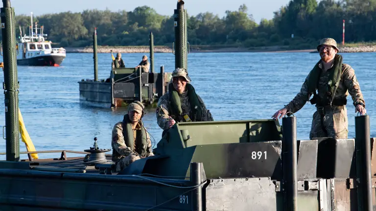 Three soldiers in camouflage uniforms and life vests operate a military boat numbered 891 on a calm river.