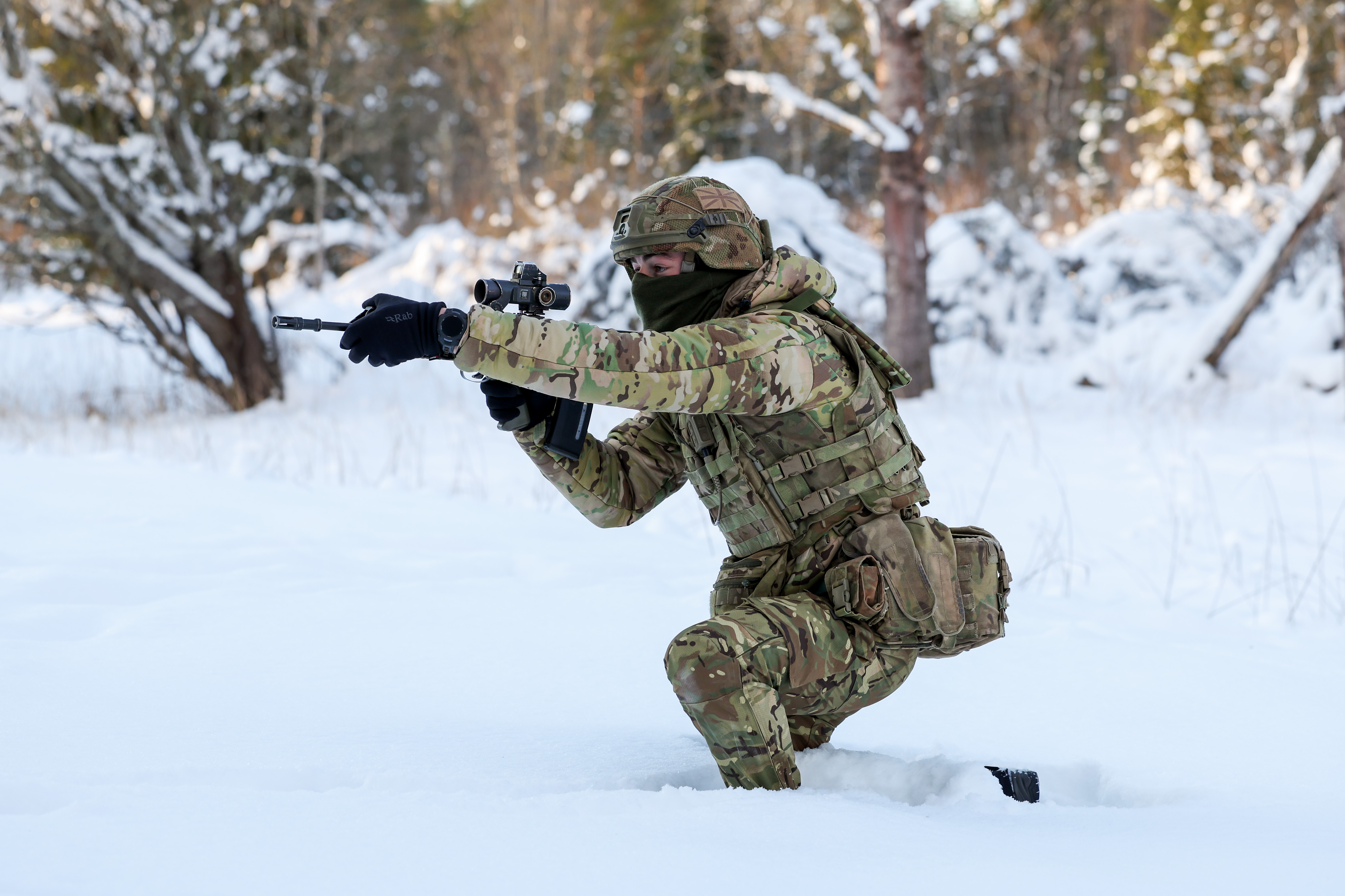 Soldier in full camouflage gear aiming a rifle while kneeling in deep snow during a winter forest training exercise.