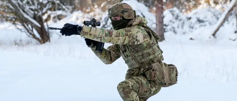 Soldier in full camouflage gear aiming a rifle while kneeling in deep snow during a winter forest training exercise.