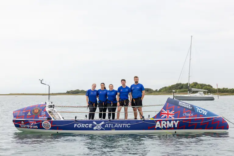 Five rowers in matching blue shirts stand on a sleek rowing boat labeled Force Atlantic Army on calm water.